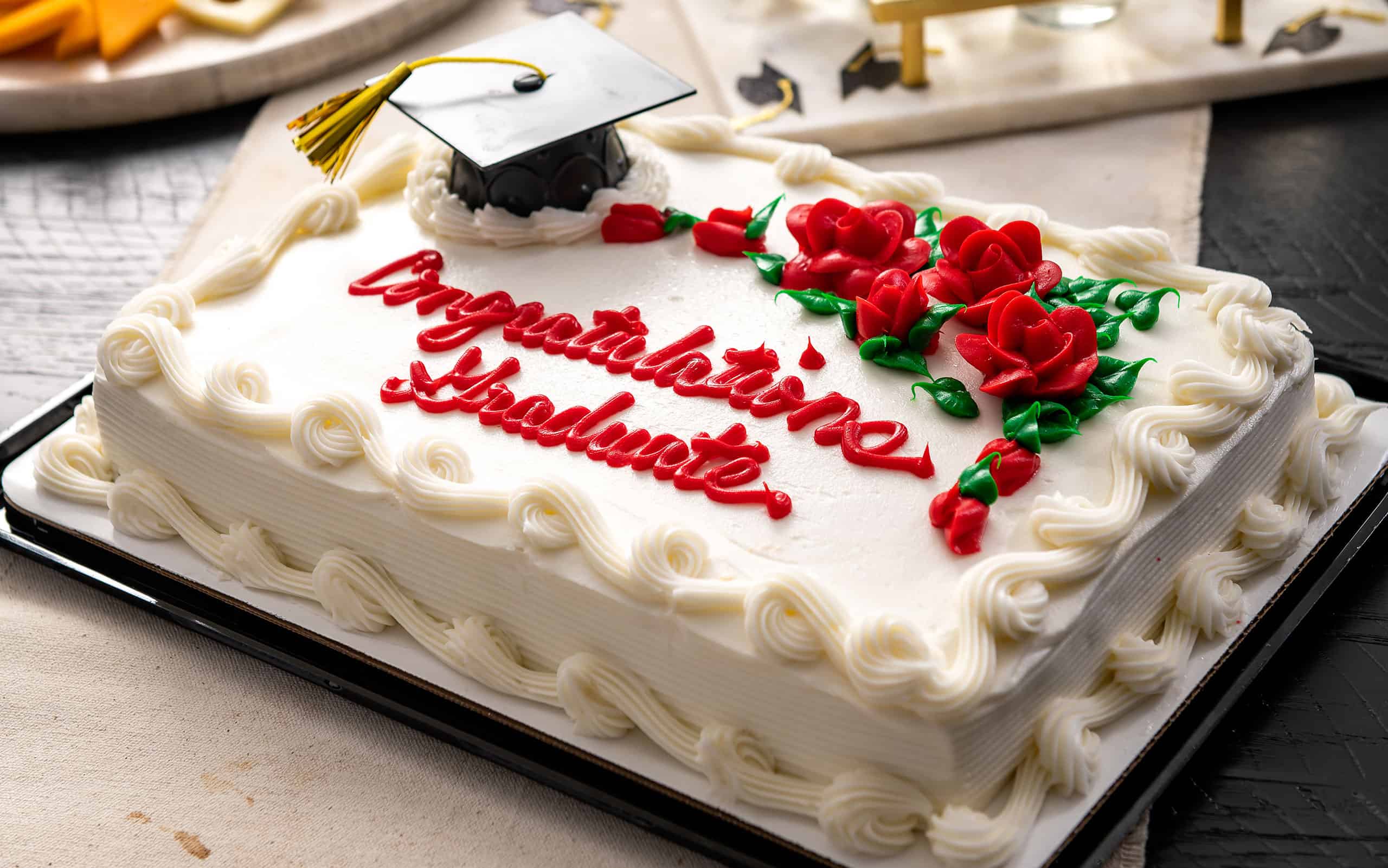 Graduation sheet cake with white frosting, red piped "Congratulations Graduate" text, red frosting roses, and a miniature black graduation cap topper.
